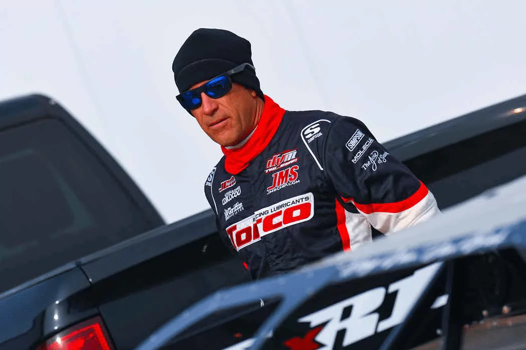Man in racing suit and sunglasses near a race car.